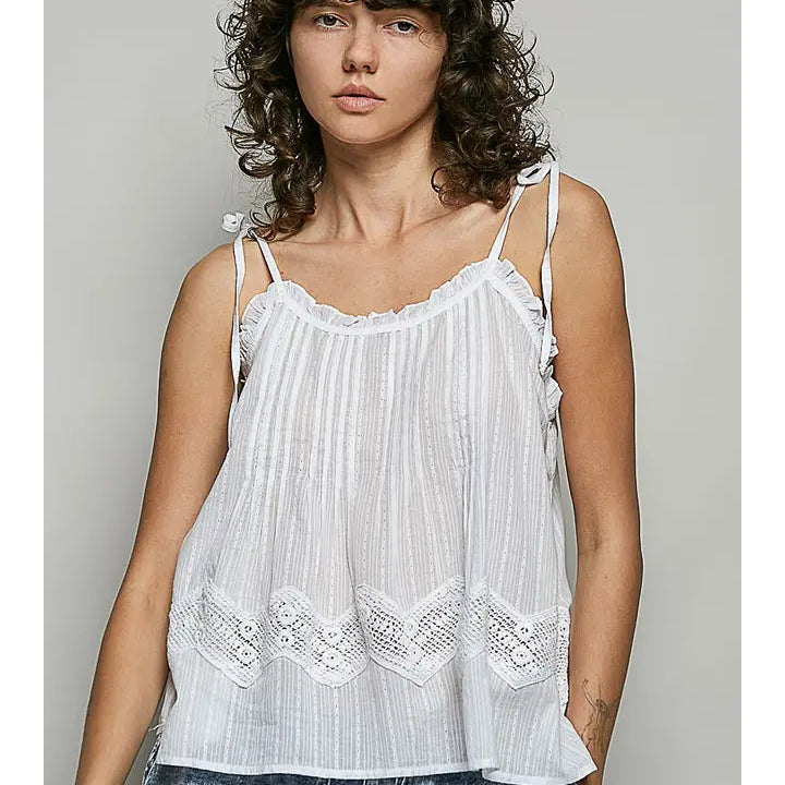 A woman wearing a white sleeveless camisole top with thin straps, ruffled trim, and embroidered lace detailing at the hem, paired with blue denim jeans against a plain gray studio background.