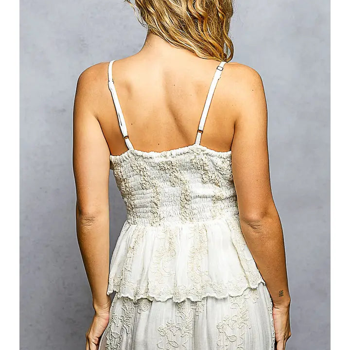 A woman shown from the back wearing a white tiered spaghetti-strap dress with floral lace applique, set against a textured gray studio background.