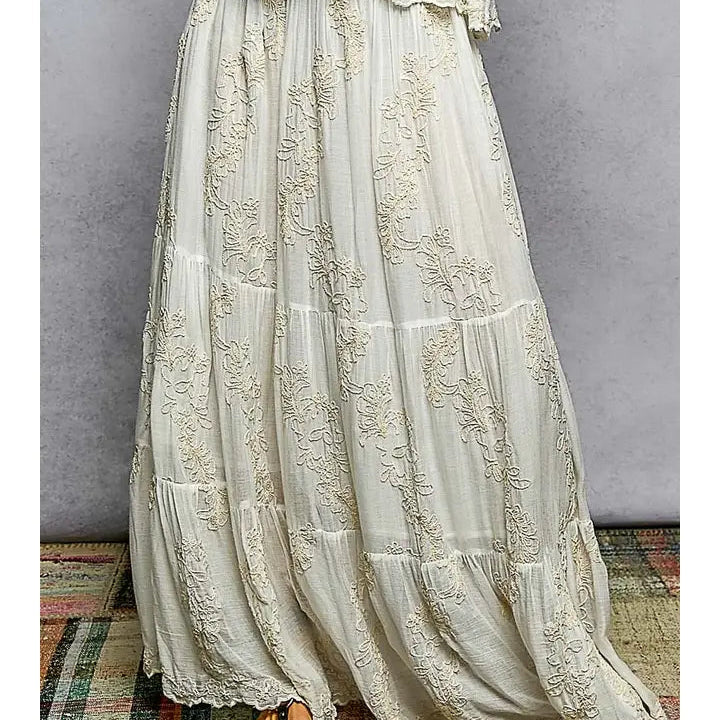 An off-white tiered maxi dress with intricate floral lace embroidery, shown on a model from the waist down against a gray textured wall.