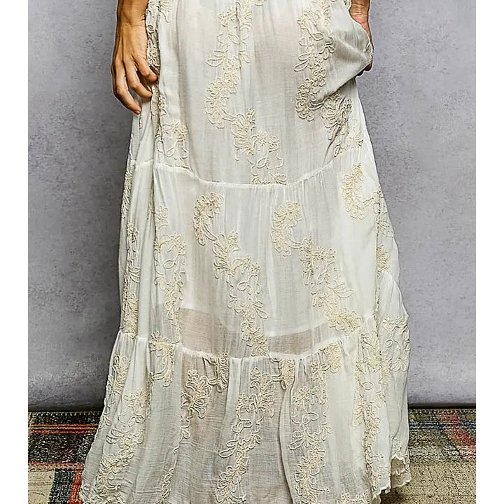 A cream-colored tiered maxi dress with intricate floral lace embroidery, shown on a model against a textured gray wall.
