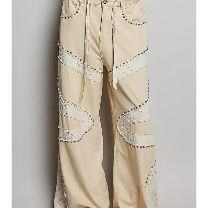 Beige wide-leg pants with cream lace applique patches and silver-tone stud detailing, shown on a model from the waist down against a light gray background.
