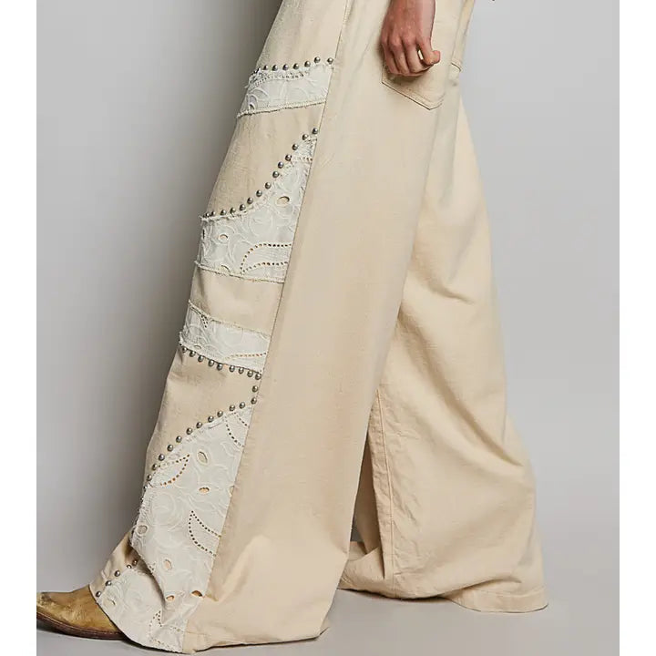 A person wearing wide-leg beige pants with decorative embroidered panels and metallic stud detailing, shown from the waist down against a light gray studio background.