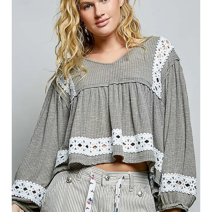 A woman modeling a gray and white striped long-sleeve crop top with white lace trim, paired with matching striped pants featuring a red, white, and blue patchwork pocket design, against a solid gray studio background.