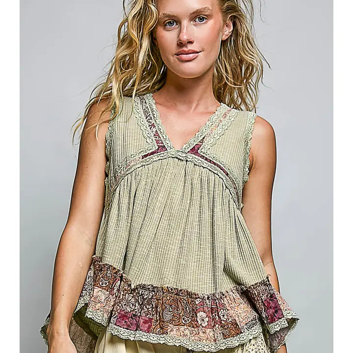 A woman wearing a sleeveless bohemian-style top with a beige and green gingham body, a multicolor paisley patterned ruffle hem, and lace trim details, paired with cream-colored pants against a solid gray studio background.
