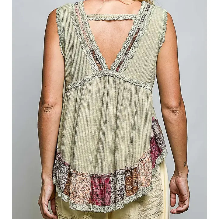 A woman shown from the back wearing a sleeveless, light beige textured top with a lace-trimmed open V-back and a ruffled multicolor patterned hem, paired with light beige pants against a neutral gray background.
