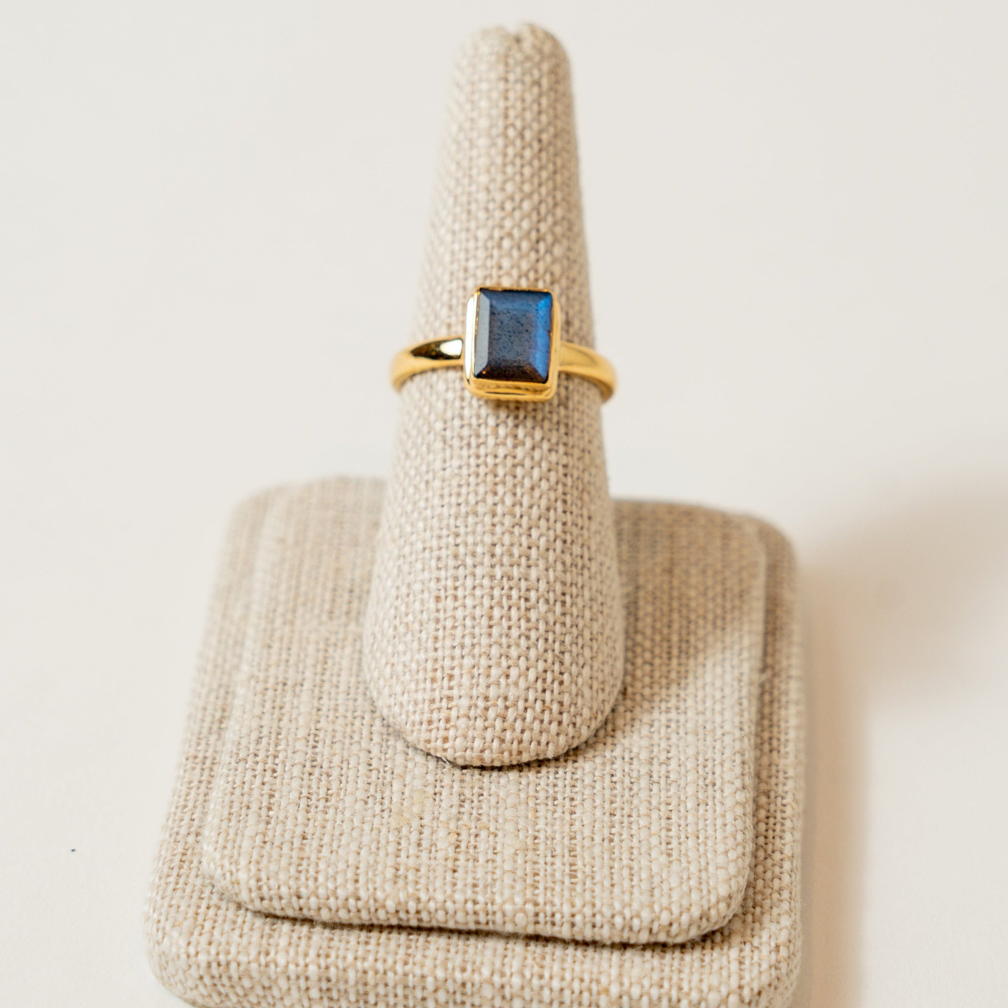 Gold ring with a blue Labradortite gemstone on a beige textured stand against a white background
