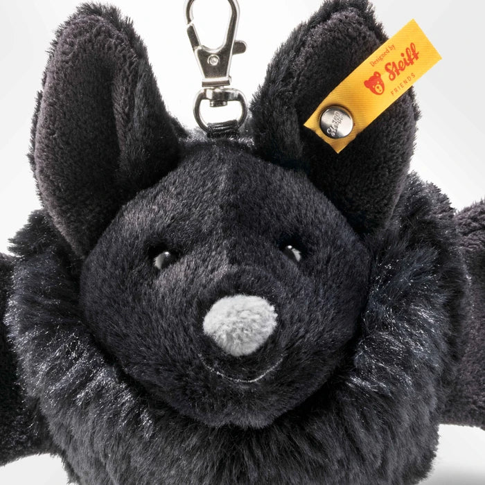 A close-up of a black plush bat keychain with a metal clasp and a yellow Stoll feather tag on a white background.