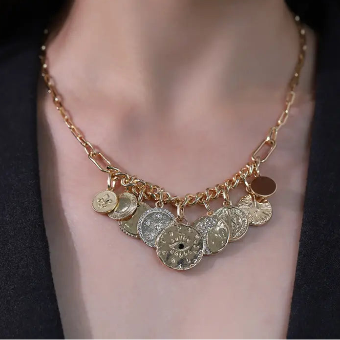 A gold-toned chain necklace with multiple dangling coin charms, worn on a model's neck against a dark blazer.