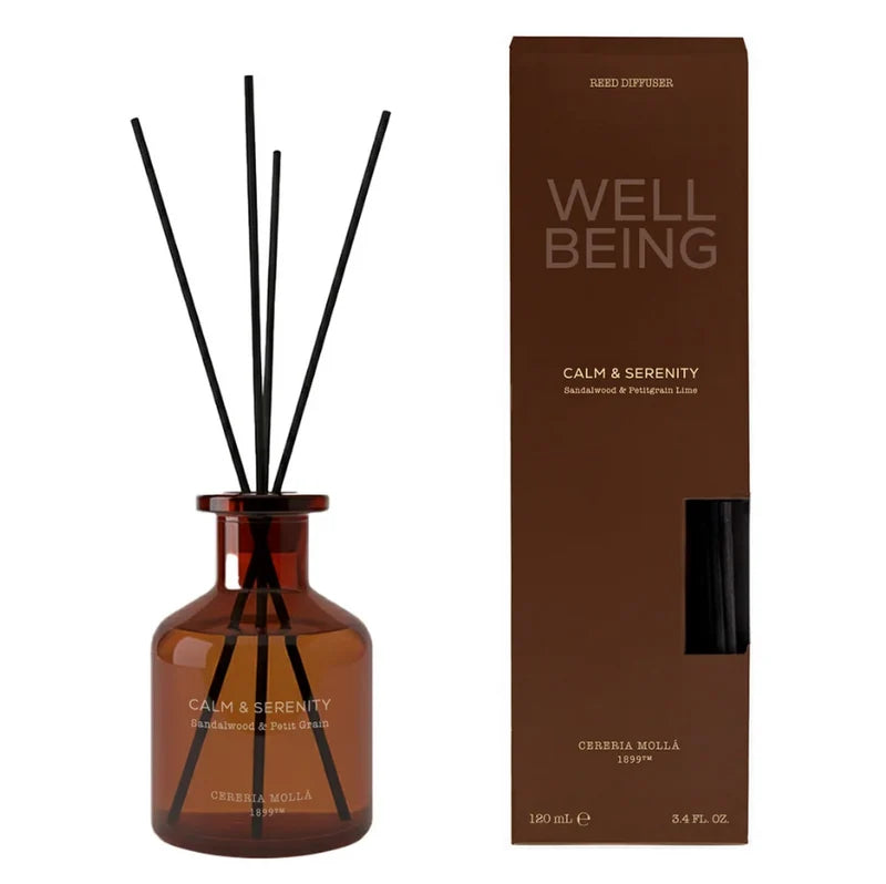 An amber glass reed diffuser bottle with black reeds next to its brown packaging box, labeled 'WELL BEING' and 'CALM & SERENITY' by Ceremia Molla.
