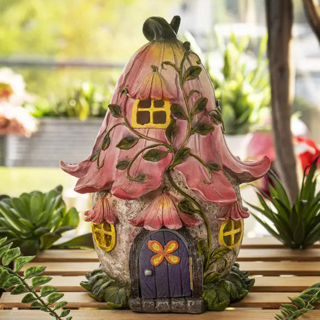 Decorative fairy house with pink flower roof on a wooden surface with plants.