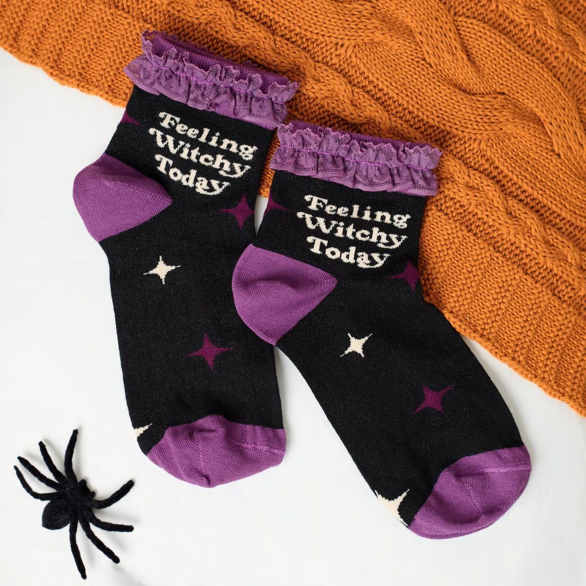 A pair of black and purple ruffled socks with 'Feeling Witchy Today' printed in white, styled on a white surface with an orange cable-knit blanket and a small black spider toy.