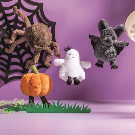 Three plush Halloween toys—a brown spider on a black web, a white ghost, and a gray bat—along with a small orange pumpkin on grass, staged against a purple background with a moon cutout.