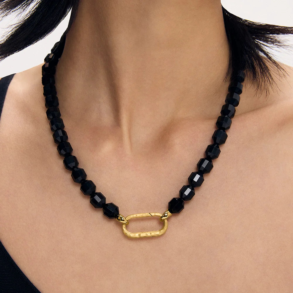 A close-up of a model's neck and chest wearing a black faceted obsidian bead necklace with a textured gold oval clasp.