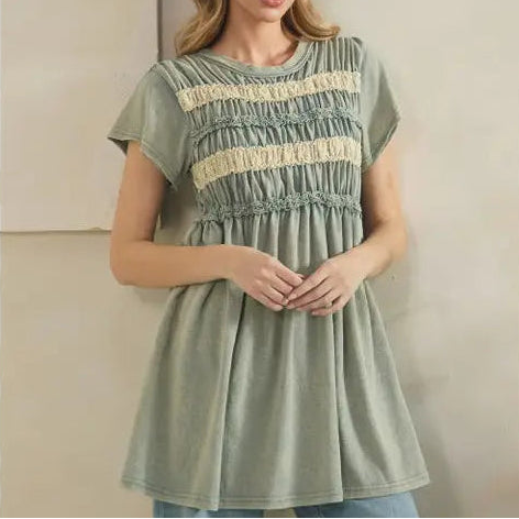 A woman wearing a sage green short-sleeve tunic top with cream horizontal lace stripes and smocked detailing, paired with light blue jeans, standing against a neutral beige wall.
