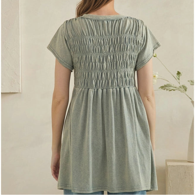 A woman viewed from the back wearing a sage green smocked short-sleeve tunic top and light blue jeans in a minimalist indoor setting.