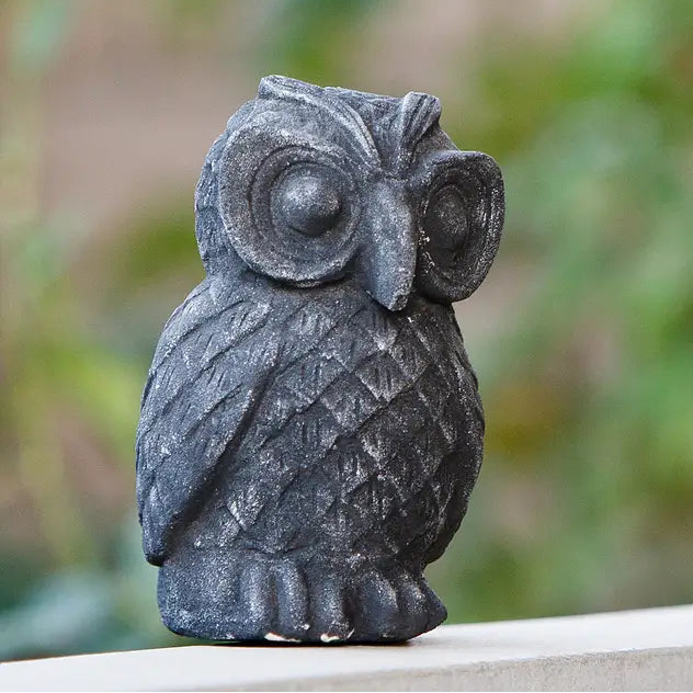 A small dark gray owl figurine with a textured stone-like finish, standing on a light-colored ledge with a blurred green and brown outdoor background.