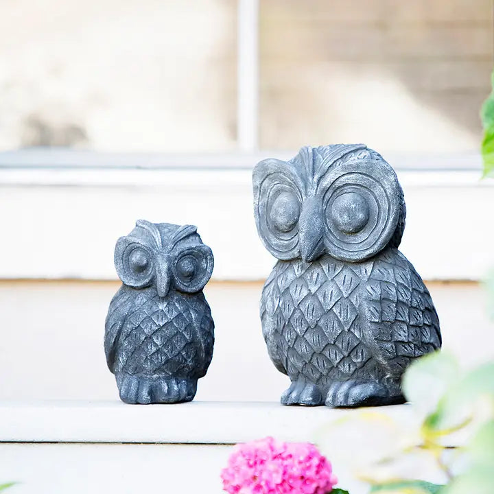 Two dark gray owl garden statues in different sizes, styled on a light-colored stone ledge with a soft-focus background and a pink flower in the foreground.