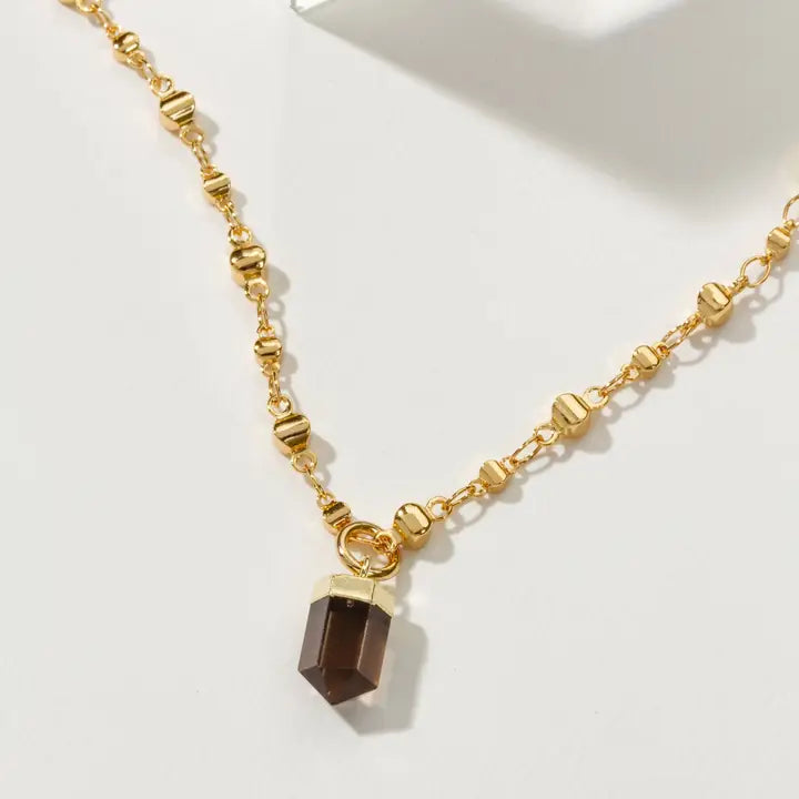 A gold-toned beaded chain necklace with a faceted smoky quartz crystal pendant, displayed on a clean white surface.
