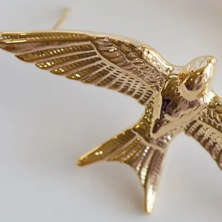 A close-up of a gold-toned bird pendant or brooch with spread wings, shown against a plain light background.