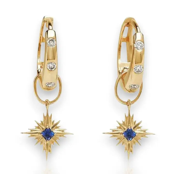 A pair of gold huggie hoop earrings with small clear stones and a dangling starburst charm featuring a blue sapphire gemstone in the center, shown on a white background.