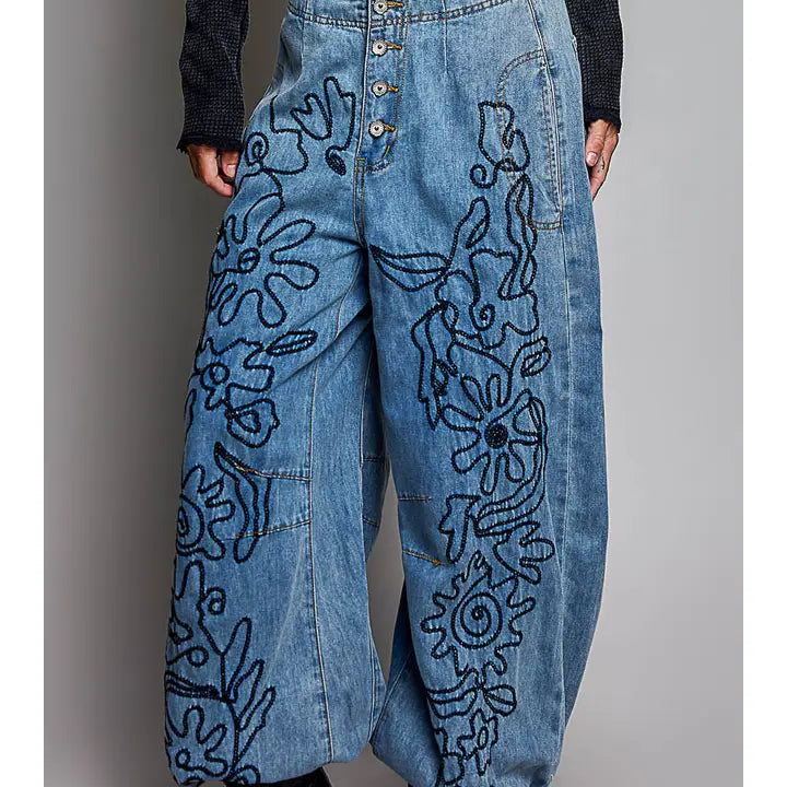 A person wearing high-waisted wide-leg blue denim pants with large black floral embroidery, paired with a dark long-sleeve top and black heeled boots, shown against a plain gray studio background.