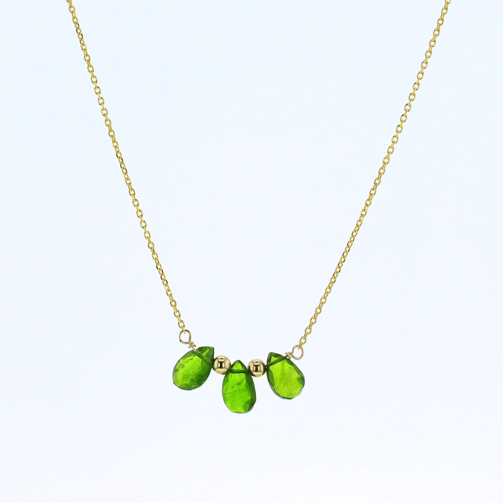 A delicate gold chain necklace featuring three faceted green tourmaline gemstone beads in a row on a light blue background.