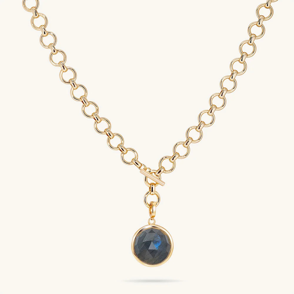 A gold chain necklace with a toggle clasp and a round bezel-set dark labradorite stone pendant, shown on a light cream background.