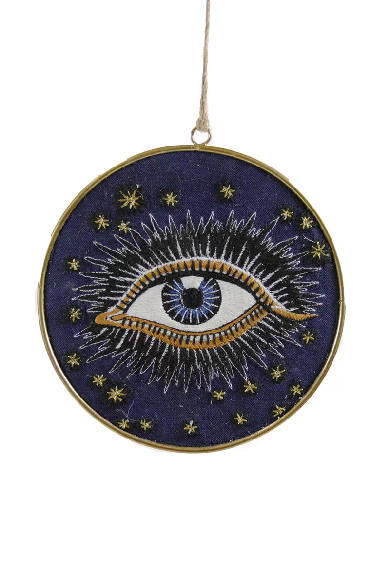 Embroidered evil eye design on a blue background with gold accents, hanging from a string.
