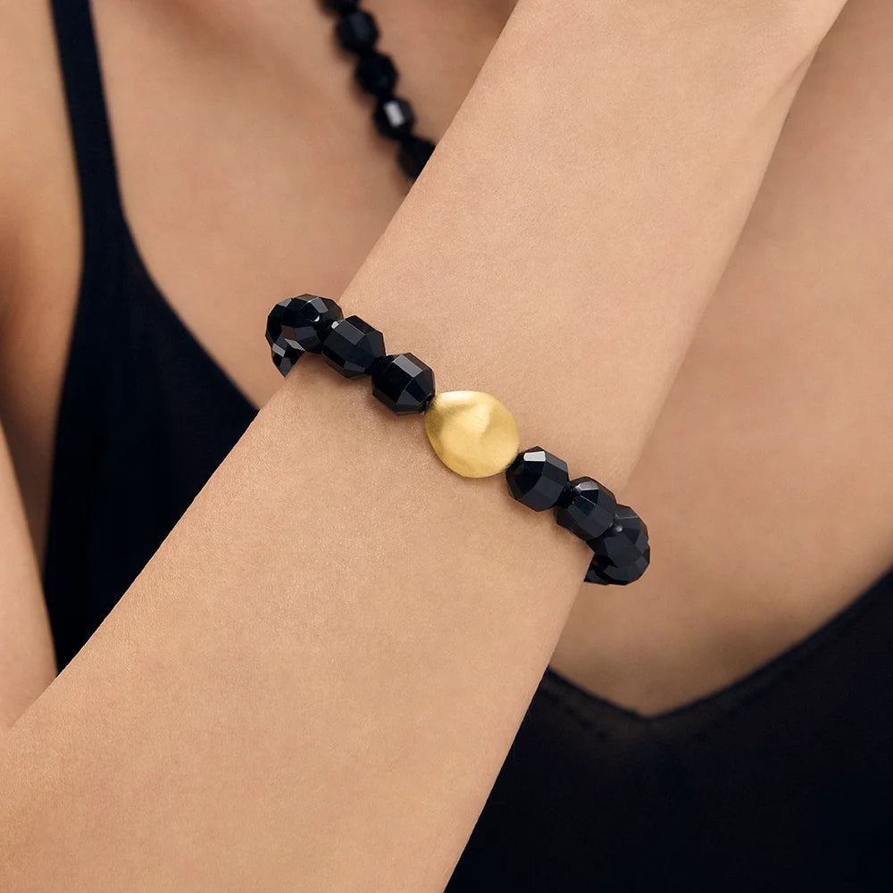 A close-up of a beaded bracelet worn on a person's wrist. The bracelet features faceted black beads and a single smooth, polished gold oval bead at the center.