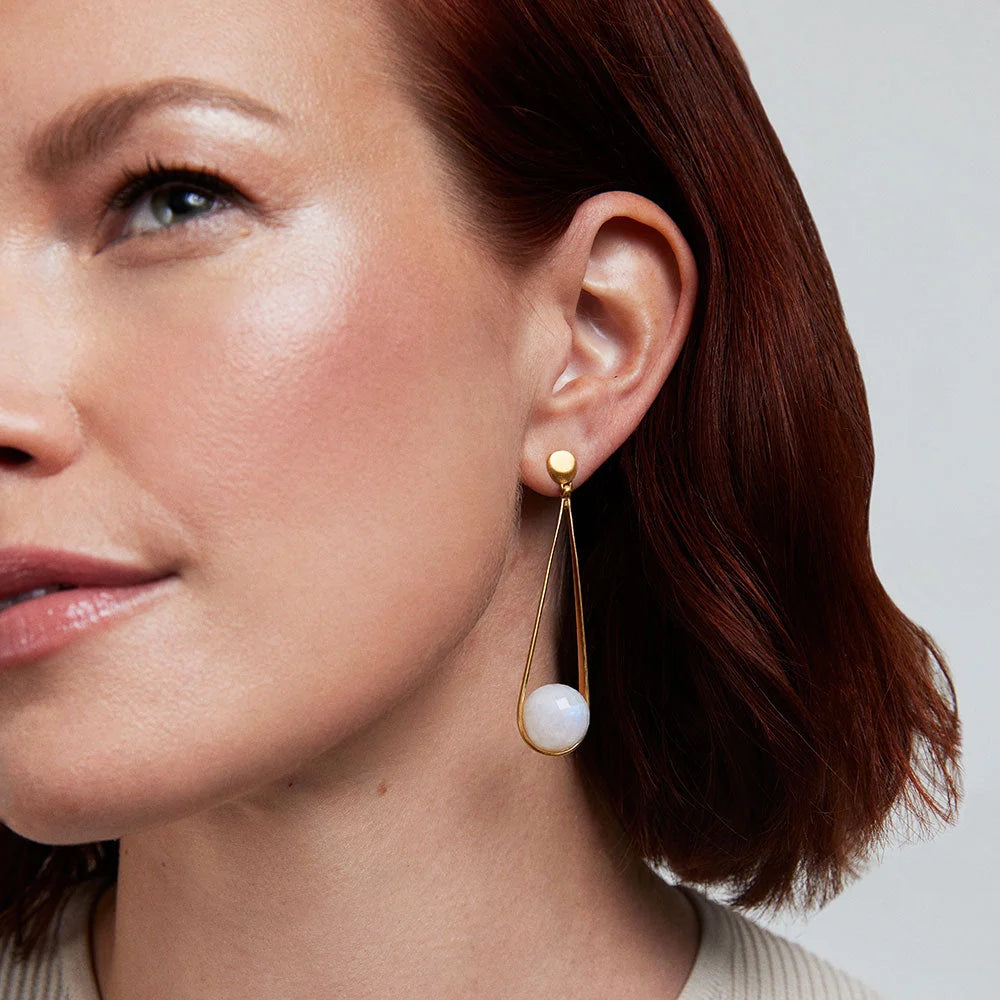 Close-up of a woman wearing gold earrings with a white moonstone on a neutral background