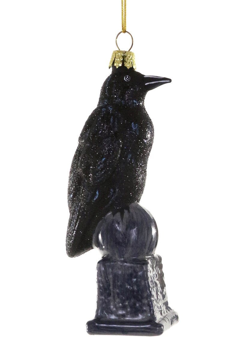 A black raven Christmas ornament with a glittery finish, perched on a dark pedestal base with a gold hanging loop at the top, shown against a plain white background.