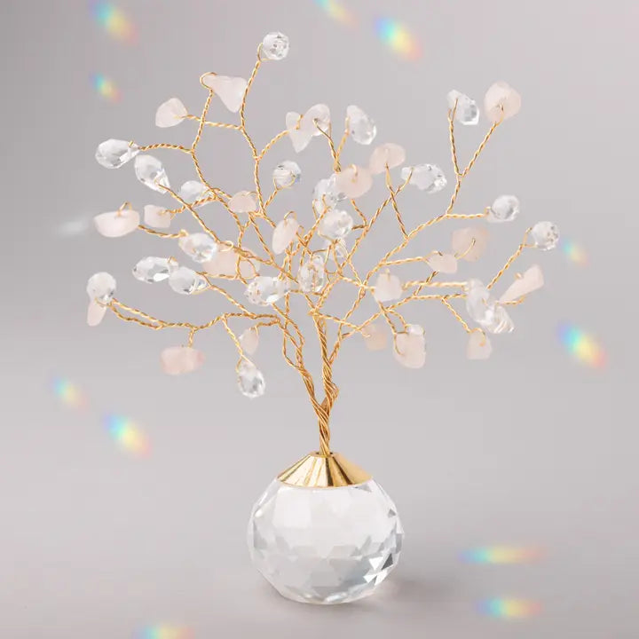 A decorative crystal tree centerpiece with gold-tone twisted branches, clear faceted crystal drops, and pale blush pink stone leaves, mounted on a clear faceted crystal sphere base against a soft gray background with subtle rainbow light leaks.
