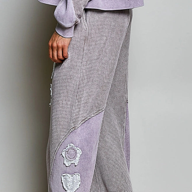 A person wearing wide-leg lounge pants in a light gray and white houndstooth pattern with lavender-colored contrast panels and white embroidered floral details down the leg, paired with black flip-flops.