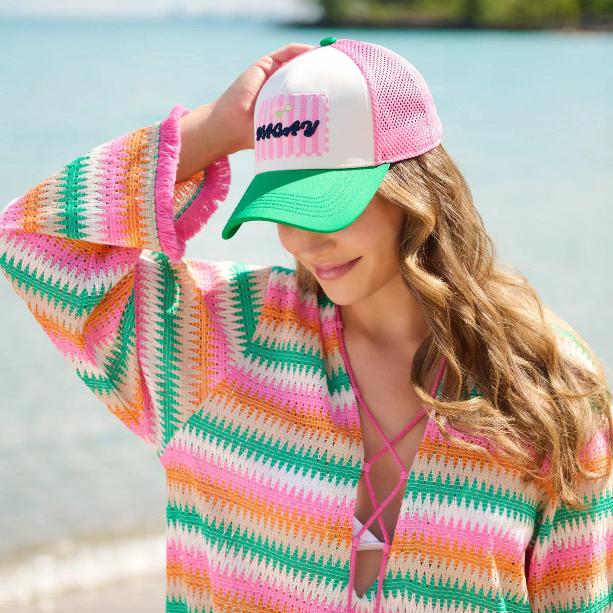 Woman wearing a colorful striped cover-up and a pink and green cap by a body of water.