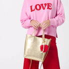 Person wearing a pink sweatshirt holding a gold tote bag with red heart keychain charm