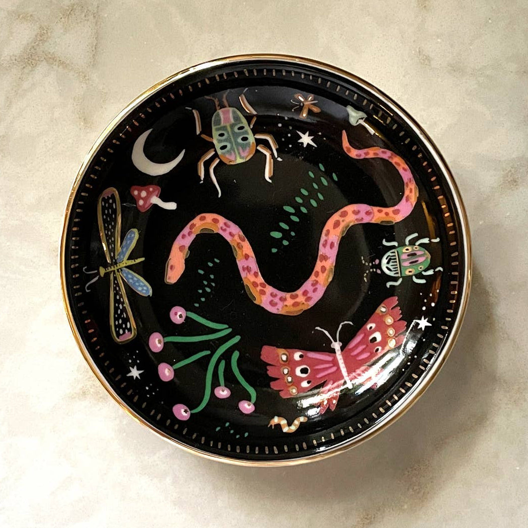 A round black decorative plate with a gold rim, featuring whimsical painted illustrations of a pink snake, butterfly, dragonfly, insects, stars, and a crescent moon, photographed from above on a light marble surface.