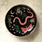 A round black decorative plate with a gold rim, featuring whimsical painted illustrations of a pink snake, butterfly, dragonfly, insects, stars, and a crescent moon, photographed from above on a light marble surface.