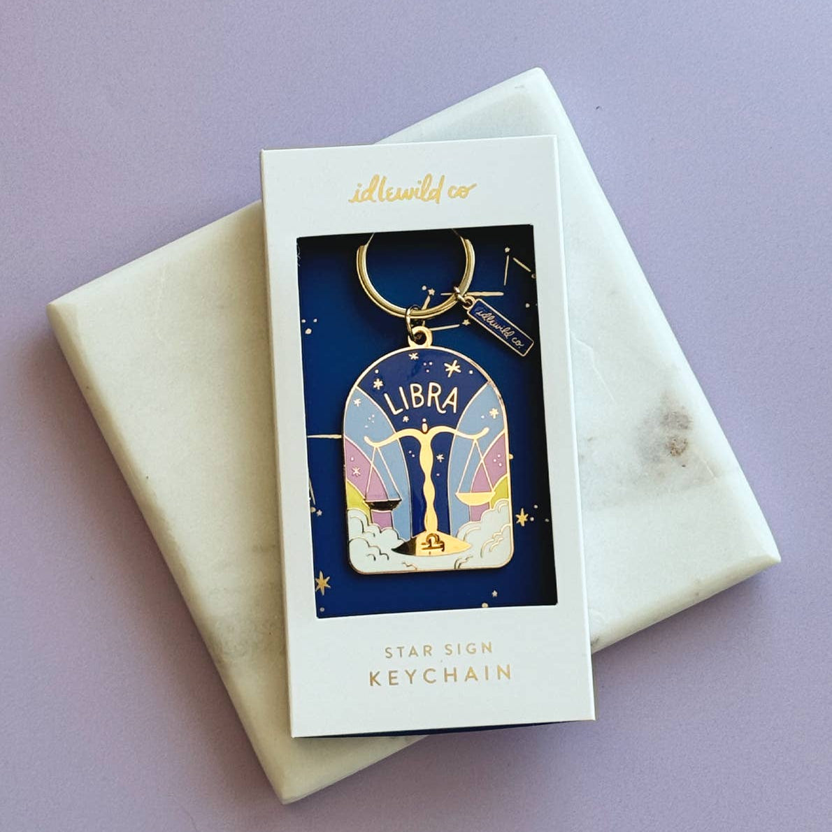 A Libra star sign keychain by iddworld.co, displayed in its original white window box packaging on a cream marble envelope against a lavender background.