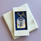 A Libra star sign keychain by iddworld.co, displayed in its original white window box packaging on a cream marble envelope against a lavender background.