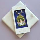 An enamel star sign keychain by Alchemy & Co, featuring a celestial design with a figure under a starry sky, presented in its original white window box packaging on a light purple background.