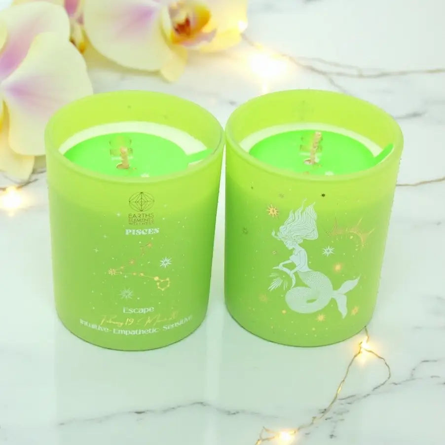 Two green Pisces Sun Sign candles with mermaid designs on a marble surface with flowers and string lights.
