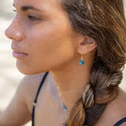 A close-up side profile of a woman wearing a small gold dangle earring with a teardrop-shaped blue flash labradorite stone, set against a soft, blurred sandy background.