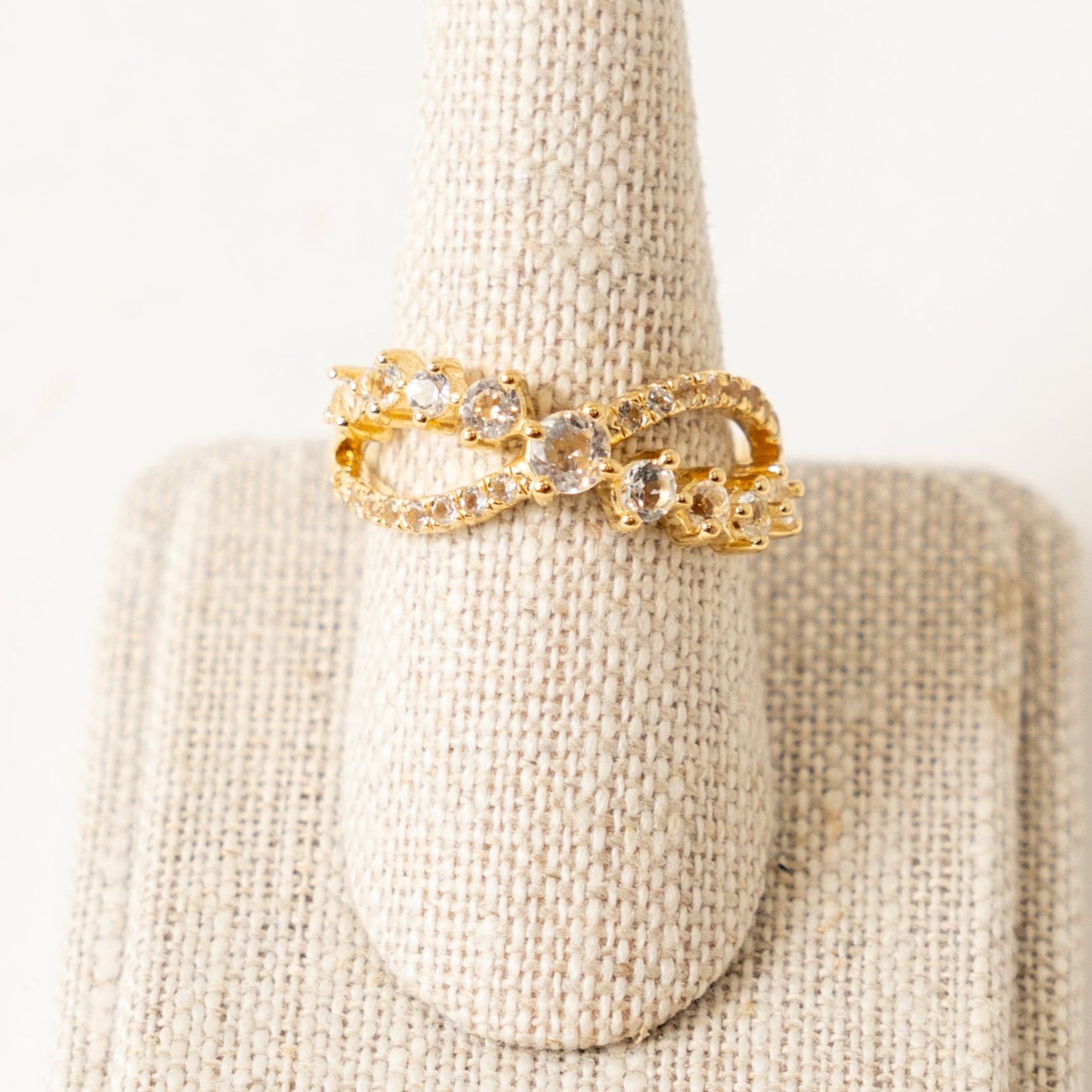 Gold ring with white topaz crystals on a beige textured stand against a white background