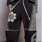 A person wearing loose black patchwork pants with gray floral and textured side panels, a matching dark top, and brown sandals, standing against a gray wall on a multicolored rug.