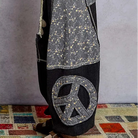 A person wearing a long, loose patchwork garment in dark gray and black with a small floral print, a large peace sign patch on the leg, and brown sandals, standing on a patterned rug against a gray wall.