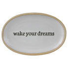 An oval white ceramic tray with a natural beige rim and the phrase 'wake your dreams' printed in black lowercase serif text across the center, shown on a plain white background.