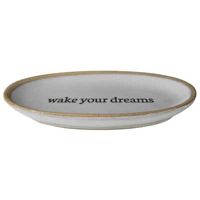 An oval ceramic dish with a rustic white glaze, a natural beige rim, and the phrase 'wake your dreams' printed in black serif text across the center.