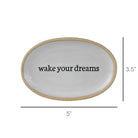 An oval white ceramic tray with a natural beige rim and the phrase 'wake your dreams' printed in black in the center. Dimension lines indicate a width of 5 inches and a height of 3.