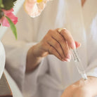 Person holding a dropper with a clear liquid, wearing a white robe with flowers in the background.