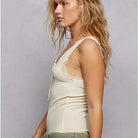 A woman in profile wearing a cream-colored lace-trimmed camisole top and green patchwork shorts against a textured gray studio background.